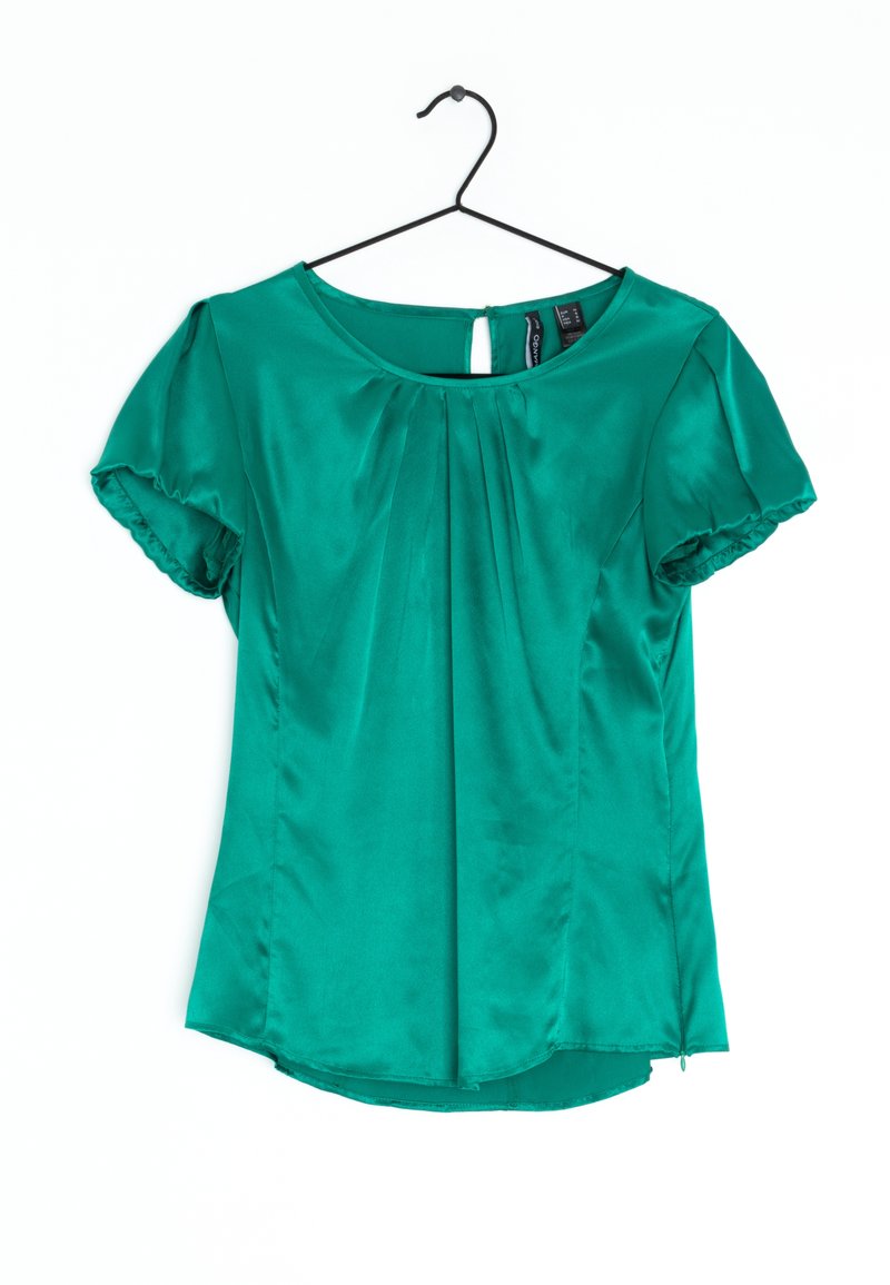 Blouse verte en satin à manches courtes avec col rond, plis avant et fermeture par bouton à ouverture en forme de goutte, suspendue à un cintre noir sur fond blanc.