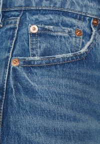 Bolsillo de jeans de mezclilla azul con remaches de cobre y costuras amarillas visibles, mostrando la textura de la tela y ligeros detalles de desgaste.