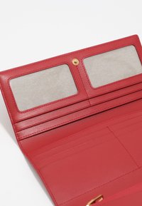 Czerwony skórzany portfel wewnątrz z dwoma przezroczystymi slotami na identyfikatory, sześcioma slotami na karty i większą kieszenią. Złote detale na zapięciu.
