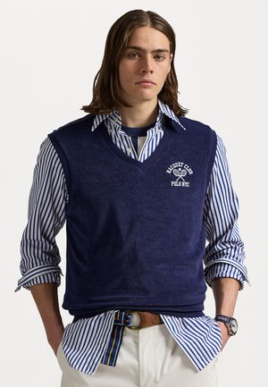 Gilet di maglia blu navy sopra una camicia a righe blu e bianche, con un logo di racchetta da tennis e una cintura marrone con fibbia rotonda.