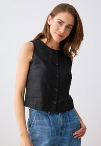 Jeune femme portant un haut noir sans manches avec des boutons et un jean bleu taille haute, posant devant un fond clair uni.