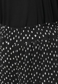 Černá skládací sukně se smockovaným páskem, která má bílý puntíkovaný vzor. Hladká textura, s různou velikostí a umístěním puntíků.