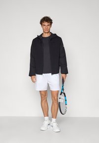 Veste rembourrée noire avec capuche, portée sur un haut sombre. Le modèle tient une raquette de tennis, portant un short blanc et des baskets blanches.