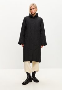 Sort, lang pufferjakke med høj krave, sidelommer og et strømlinet design. Båret over cremefarvede tapered bukser og sorte støvler.