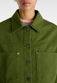 Giacca di denim verde con colletto, quattro tasche e bottoni neri. Il tessuto presenta una finitura testurizzata e dettagli di cucitura visibili.