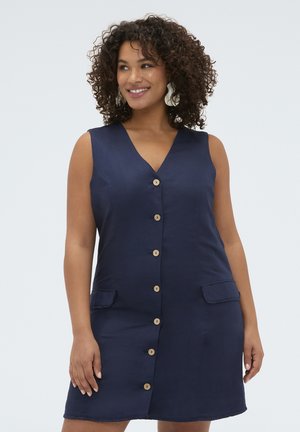 Vrouw met krullend haar, die een mouwloze navy blousejurk met knopen en voorzakken draagt, glimlachend en naar links kijkend tegen een lichte achtergrond.