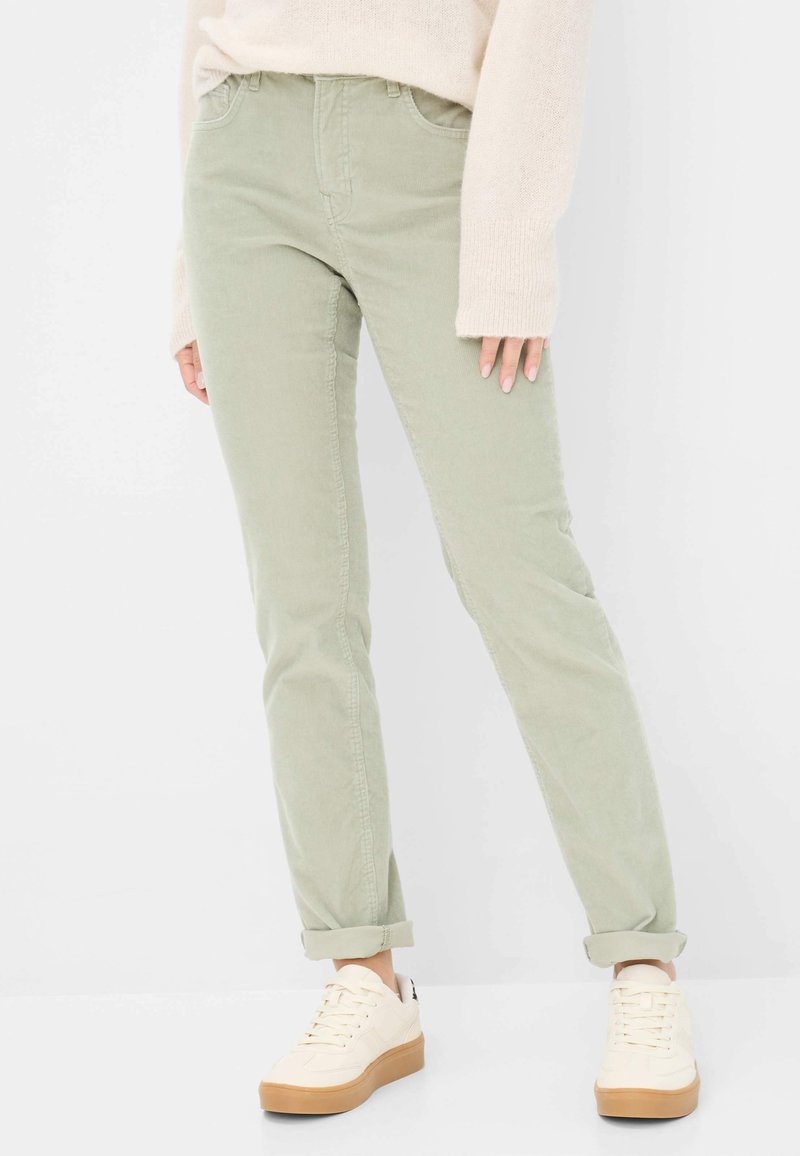 Pantaloni in corduroy verde chiaro con una vestibilità slim, dotati di tasche frontali, chiusura a zip e orli arrotolati, abbinati a sneaker bianche con suola beige.