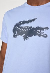Camiseta de algodão azul claro com um gráfico de crocodilo preto e padrões triangulares nas costas. Design de mangas curtas e gola redonda.