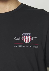 Crna pamučna majica s izvezenim grbom u crvenoj i plavoj boji, s natpisom "GANT American Sportswear" i godinom "1949."