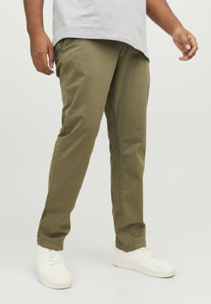 Man wearing olive green pants, white sneakers, and a light gray shirt standing against plain background.