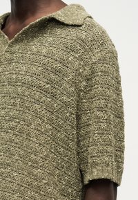 Κοντινό πλάνο ενός ατόμου με σκουρόχρωμο δέρμα που φοράει πλεκτό πουλόβερ ελιάς με υφή, φαρδιά γιακά και κοντά μανίκια.