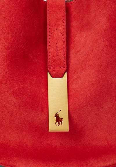 Rdeča torbica iz semiša z zlatim detajlom in logotipom v izrezu. Gladek tekstura, izrazita oblika in močna barva.