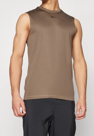 Hombre con camiseta deportiva marrón sin mangas y pantalones cortos negros, de pie con los brazos relajados contra un fondo blanco.