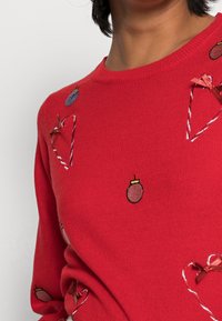 Rode gebreide trui met geborduurde feestelijke motieven, waaronder harten, ornamenten en zuurstokken, afgewerkt met strikjes en een getextureerde stof.