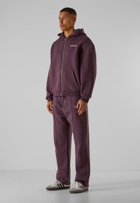 Lilla zip-up hættetrøje parret med matchende sweatpants. Lavet af blødt materiale med to forlommer og hvidt logo-detail på brystet.