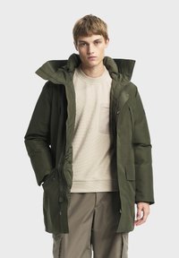 Parka à capuche vert olive avec fermeture éclair et poches, portée sur un sweat à texture beige clair et un pantalon gris clair.
