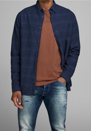 Marine gestreepte overhemd met knopen over een bruine ronde hals t-shirt, gedragen met lichtblauwe distressed spijkerbroek, die verschillende texturen en kleuren toont.