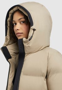 Beige Pufferjacke mit großer Kapuze, schwarzen Akzenten, Reißverschluss und gestepptem Design. Auffälliger Kordelzug zur Anpassung der Kapuze.