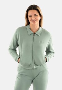 Giacca zip-up verde chiaro realizzata in tessuto morbido, con colletto, tasche laterali e iniziali ricamate "BM" sul petto.