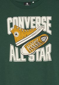 Zeleno pulover s kapuco, na sprednji strani natisnjeni rumeni čevlji Converse, z napisom "CONVERSE ALL STAR" v bold belih črkah.