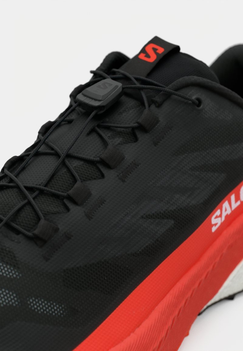 Schwarzer Laufschuh mit strukturiertem Obermaterial, schwarzen Schnürsenkeln und einer roten Außensohle mit einem weißen Logo an der Seite.