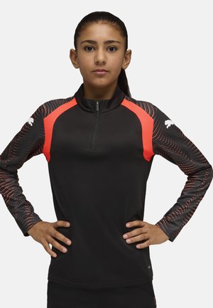 Fille aux cheveux foncés en queue de cheval, portant un haut de sport noir et rouge à manches longues avec des manches à motifs, debout les mains sur les hanches.