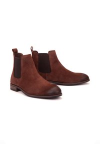 Botines de ante marrón con paneles elásticos en los laterales, punta redondeada y un tacón bajo apilado. Incluyen una lengüeta para facilitar su colocación.