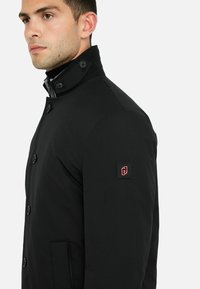 Schwarzer, knöpfbarer Mantel mit hohem Kragen und Seitentaschen; besteht aus einem strukturierten Stoff und hat ein Logo-Patch am linken Ärmel.