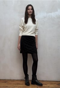 Kermanvärinen neulepusero, musta kietaisuhame hopeisilla niiteillä, mustat sukkahousut, mustat nilkkurit, seisoo puisella lattialla yksinkertaista seinää vasten.