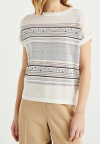 Donna che indossa un top bianco a maglia a maniche corte con motivi geometrici orizzontali neri e beige e pantaloni beige a vita alta.
