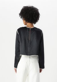 Cropped black top with long sleeves, white stitching, and a back zipper detail; paired with white high-waisted pants.