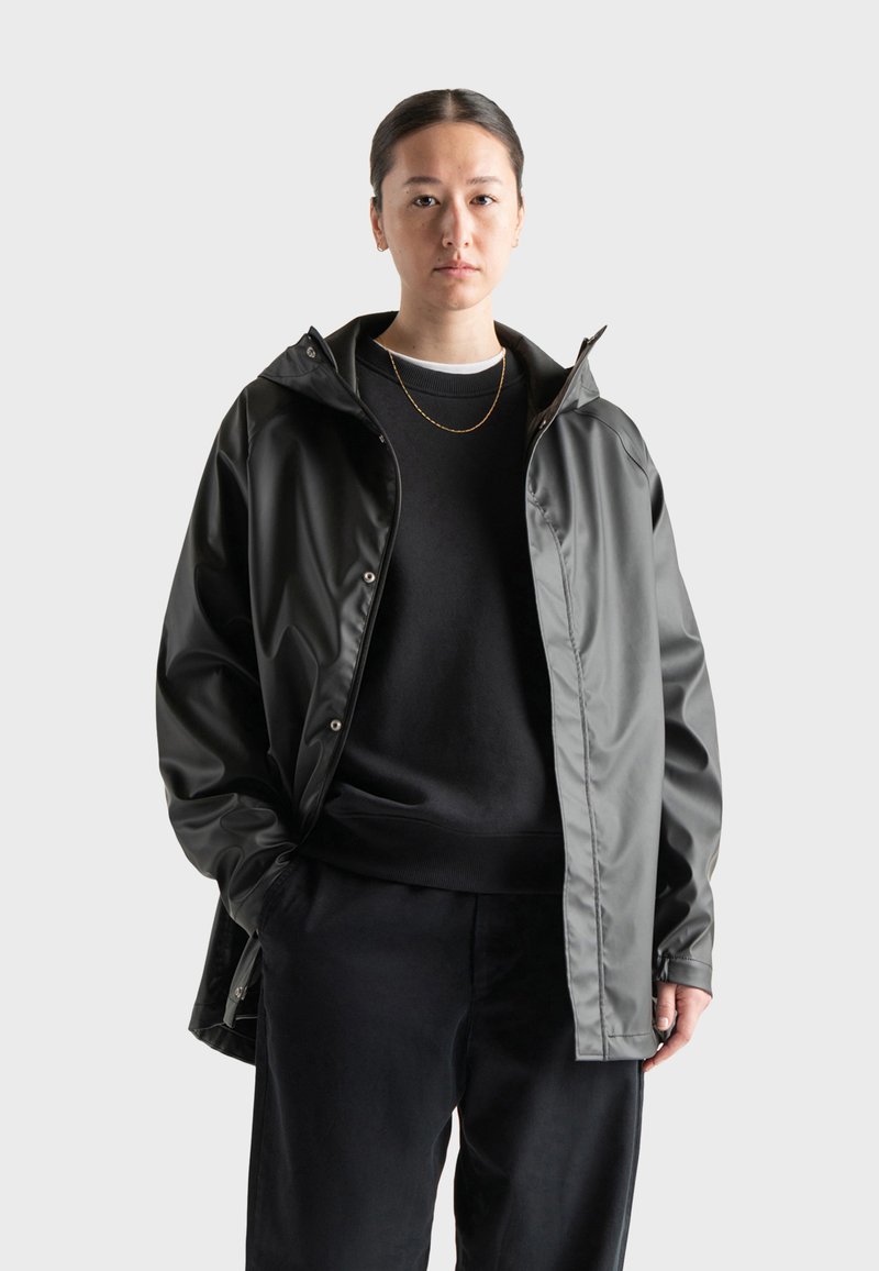 Chaqueta impermeable en negro y gris con capucha, cierres a presión y un corte relajado. Lleva puesta sobre una sudadera negra y pantalones.