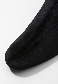 Gros plan sur l'avant d'une chaussure en tissu noir avec des coutures visibles sur fond blanc.