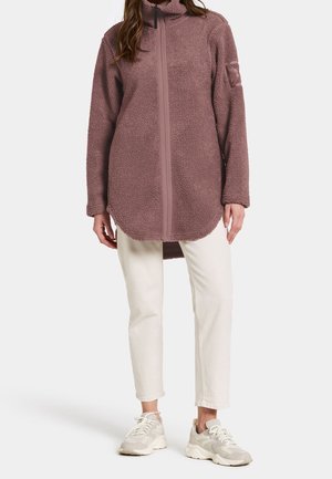Kvinde iført støvet rosa oversized fleecetrøje med frontlynlås, hvide cropped bukser og hvide sneakers, stående mod ensfarvet baggrund.
