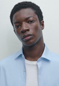 Lichtblauw button-up shirt over een wit T-shirt. Het shirt heeft een klassieke kraag en is gemaakt van een gladde stof. Close-up van het bovenlichaam.