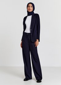 Abito in tonachina navy con pantaloni lunghi e larghi e giacca su misura. Top bianco aderente sotto. I dettagli includono due bottoni sulla giacca.