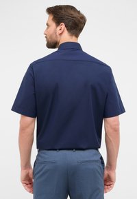 Mann mit braunen Haaren und Bart, der ein dunkelblaues Kurzarmhemd und hellblaue Hosen trägt, mit dem Rücken zu einer schlichten weißen Hintergrund.