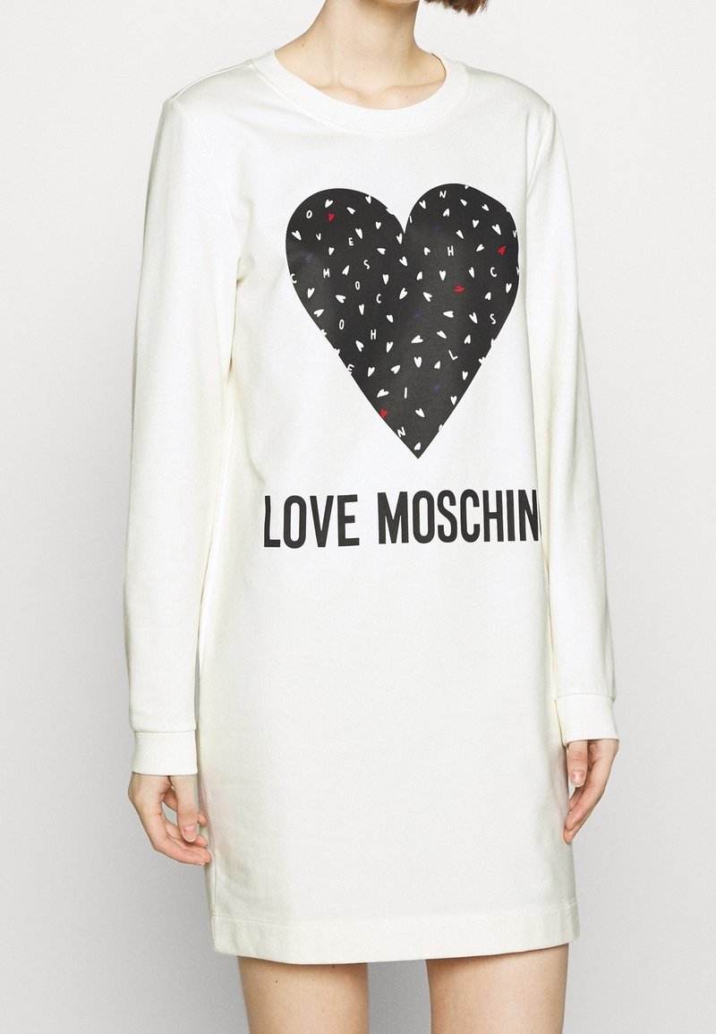 Bílé šaty s dlouhým rukávem, které mají černou srdcovou grafiku s barevnými akcenty a textem "LOVE MOSCHINO" v tučném písmu na spodní části.