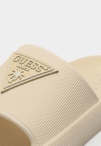 Gros plan sur la bride beige de la sandale slide Guess Beach, avec une surface texturée et un logo triangulaire représentant un palmier.