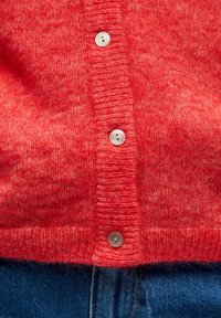 Sarkana adīta kardigāna ar teksturētu virsmu, pieciem gaišiem pogām un ribu apmali. Kombinēta ar zilu džinsu audumu.