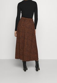 Persoon in een zwart langemouwen top, een midirok met een bruine en zwarte print, en zwarte enkellaarsjes met hak, staand voor een effen achtergrond.