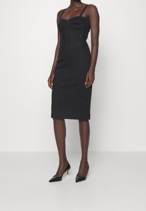 Femme portant une robe noire moulante longueur genou avec fines bretelles et des escarpins noirs pointus avec bouts de talons vert fluo.