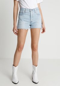 Shorts de mezclilla azul claro con dobladillos deshilachados, ajuste de talle alto y bolsillos delanteros. Combinados con botas de vaquero blancas. Camiseta blanca simple visible.