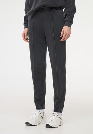 Pantaloni jogger grigio scuro realizzati in tessuto morbido, con vita elastica, cuciture pinzate e polsini affusolati, abbinati a scarpe da ginnastica bianche.