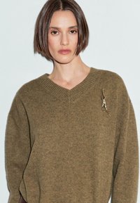 Donna con i capelli corti castani che indossa un maglione lavorato a maglia di colore verde oliva con una spilla astratta dorata sul lato sinistro.