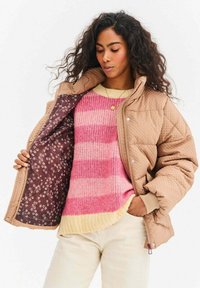 Prešívaná béžová bunda s hnedou kvetovanou podšívkou, nosená cez ružový a krémový pruhovaný sveter. Bunda má stojatý golier a zapínanie na cvoky.