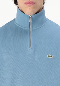 Pulover cu fermoar deschis în nuanță de albastru deschis, cu guler înalt, textură ribbed și un mic logo accent pe piept. Detaliu de fermoar argintiu la gât.