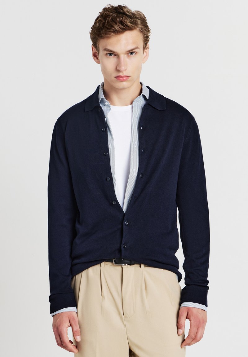 Marineblauer Cardigan mit Knöpfen, aus einem weichen Stoff. Wird über einem weißen T-Shirt und einem hellblau gestreiften Hemd getragen. Leicht und taillierte Passform.