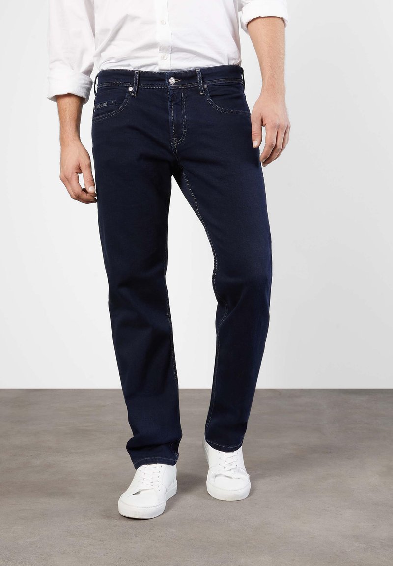 Dark blue denim jeans with a straight fit, featuring white stitching, front pockets, and a button fly, worn with white sneakers.