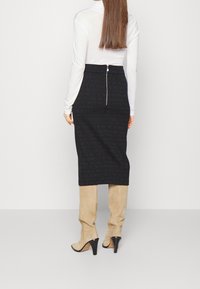 Vrouw in een witte lange mouwen top, zwarte pencilrok met een rits aan de achterkant, en beige suède hoge haklaarzen die tegen een witte muur staat.